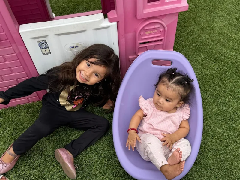 A baby sitting in the yard next to an older girl by the pink playhouse