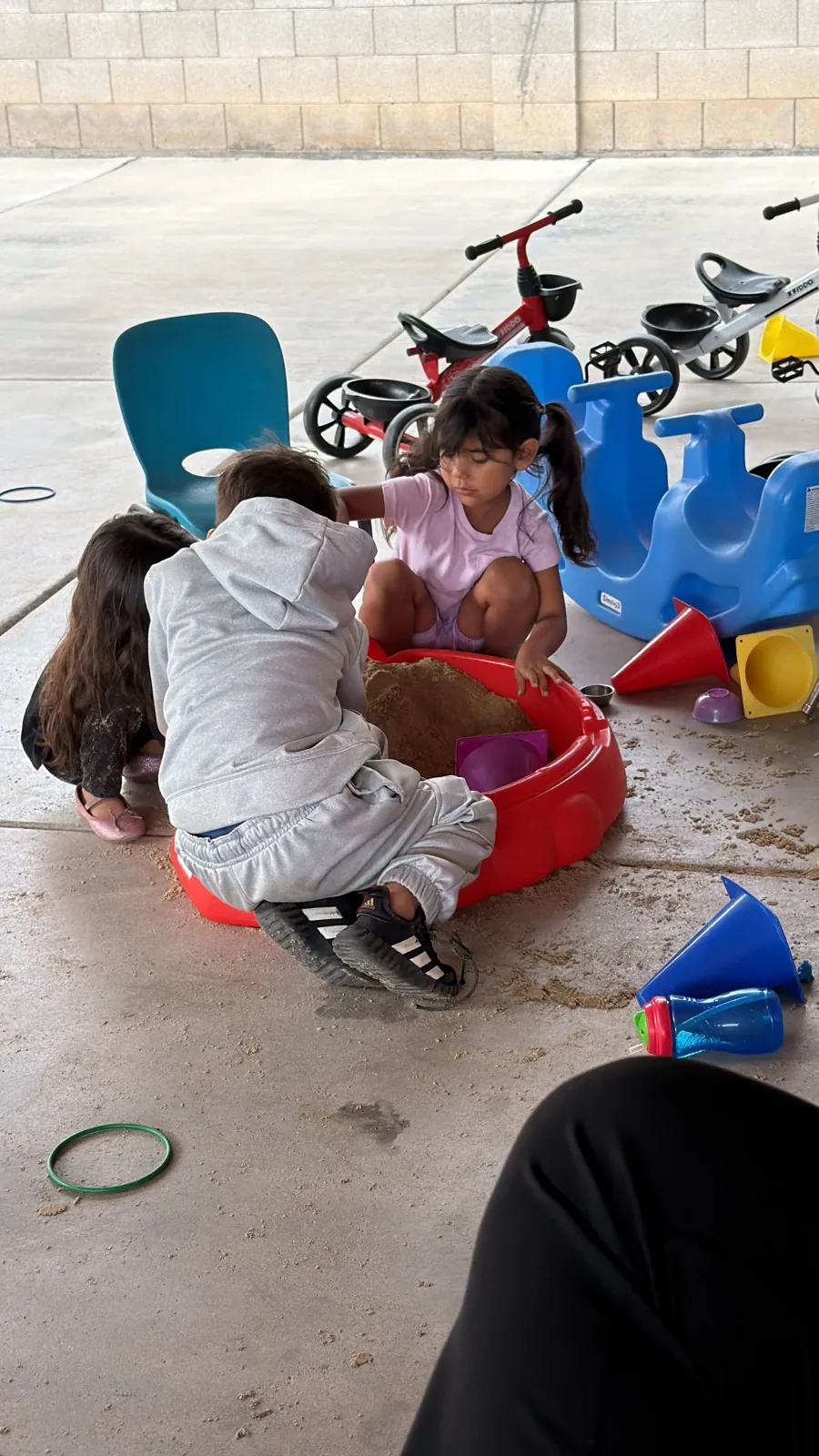Toddlers digging in the sandbox with the bikes and toys behind them